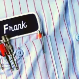 Chemise à rayures bleues et rouges avec un badge "Frank" et des stylos dans la poche. Le titre du roman est le nom du personnage principal.