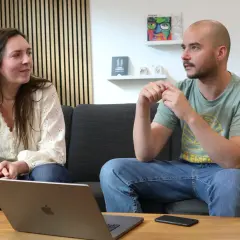 Un homme et une femme discutent sur un canapé. L'homme, chauve, porte un t-shirt vert. La femme, brune, porte une blouse blanche. Un ordinateur portable est ouvert sur la table. Un exemple de langage cru pourrait être utilisé dans leur conversation.