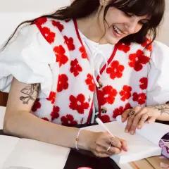 Une jeune femme souriante fait une dédicace livre, stylo à la main, entourée de cadeaux et de carnets.