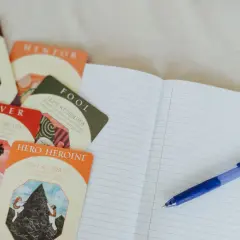 Cartes d'archétypes (Trickster, Lover, Hero/Heroine) et un carnet ouvert avec un stylo, pour une idée de nom pour personnage de roman.