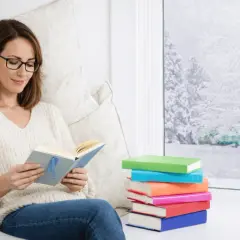 Une femme choisit un livre, confortablement installée près d'une fenêtre enneigée. Une pile de livres colorés l'attend.
