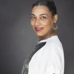 Une autrice au sourire confiant, ses cheveux courts grisonnants encadrant un visage aux taches de rousseur. Elle porte des boucles d'oreilles dorées et un t-shirt blanc.