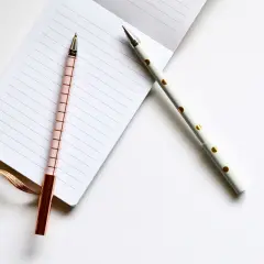 Deux stylos élégants, un rose à carreaux et un blanc à pois dorés, prêts pour l'édition pkj manuscrit sur un carnet ligné.