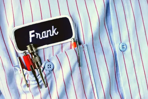 Chemise à rayures bleues et rouges avec un badge "Frank" et des stylos dans la poche. Le titre du roman est le nom du personnage principal.