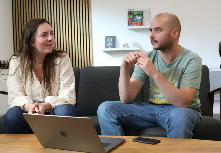 Un homme et une femme discutent sur un canapé. L'homme, chauve, porte un t-shirt vert. La femme, brune, porte une blouse blanche. Un ordinateur portable est ouvert sur la table. Un exemple de langage cru pourrait être utilisé dans leur conversation.