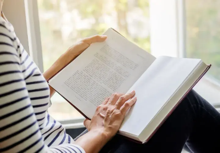 Une personne lit un livre près d'une fenêtre. Les pages sont blanches, suggérant le fin du livre.