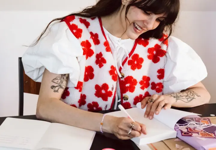 Une jeune femme souriante fait une dédicace livre, stylo à la main, entourée de cadeaux et de carnets.