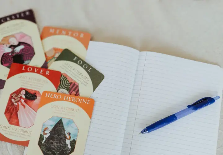 Cartes d'archétypes (Trickster, Lover, Hero/Heroine) et un carnet ouvert avec un stylo, pour une idée de nom pour personnage de roman.