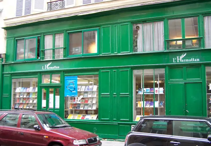 Façade verte d'une librairie L'Harmattan, vitrines remplies de livres. Une voiture rouge et une noire stationnent devant.