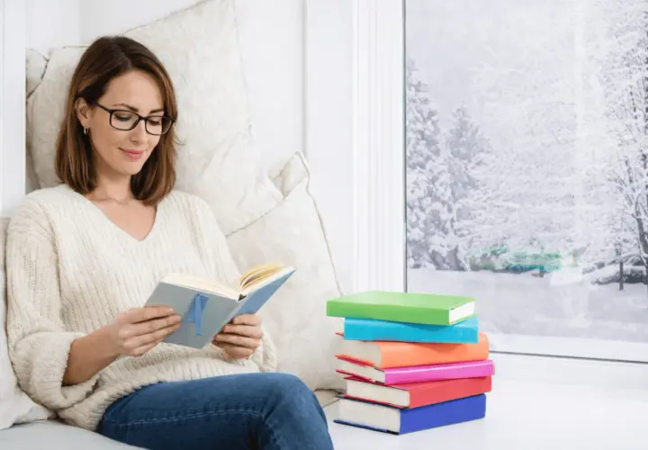 Une femme choisit un livre, confortablement installée près d'une fenêtre enneigée. Une pile de livres colorés l'attend.