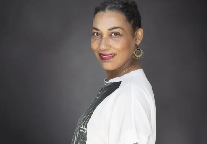 Une autrice au sourire confiant, ses cheveux courts grisonnants encadrant un visage aux taches de rousseur. Elle porte des boucles d'oreilles dorées et un t-shirt blanc.