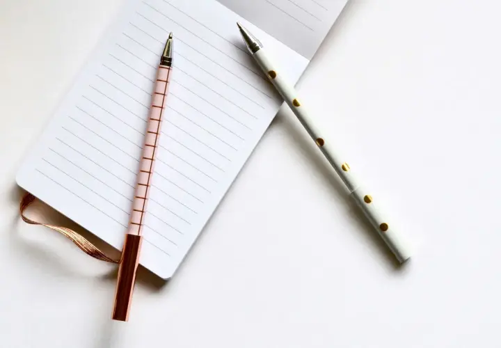 Deux stylos élégants, un rose à carreaux et un blanc à pois dorés, prêts pour l'édition pkj manuscrit sur un carnet ligné.