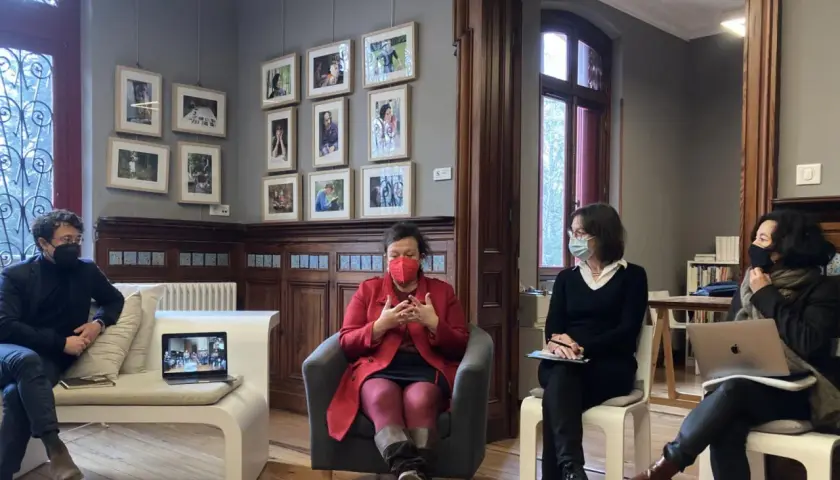 Quatre personnes masquées discutent dans un salon orné de cadres. Une femme en rouge est au centre, semblant raconter une histoire lors d'une résidence d'écriture.
