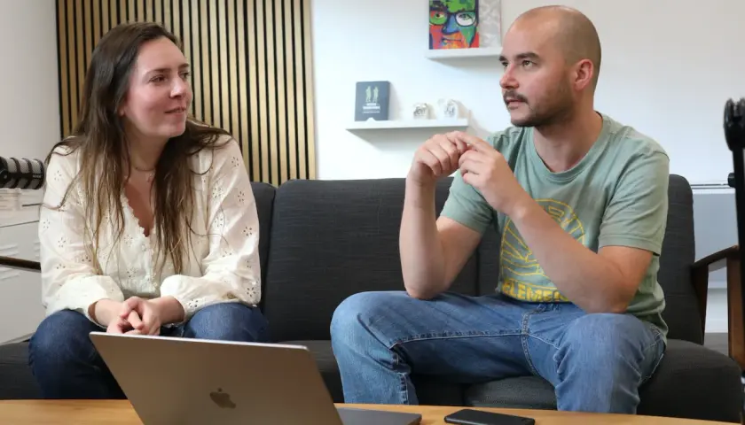 Un homme et une femme discutent sur un canapé. L'homme, chauve, porte un t-shirt vert. La femme, brune, porte une blouse blanche. Un ordinateur portable est ouvert sur la table. Un exemple de langage cru pourrait être utilisé dans leur conversation.
