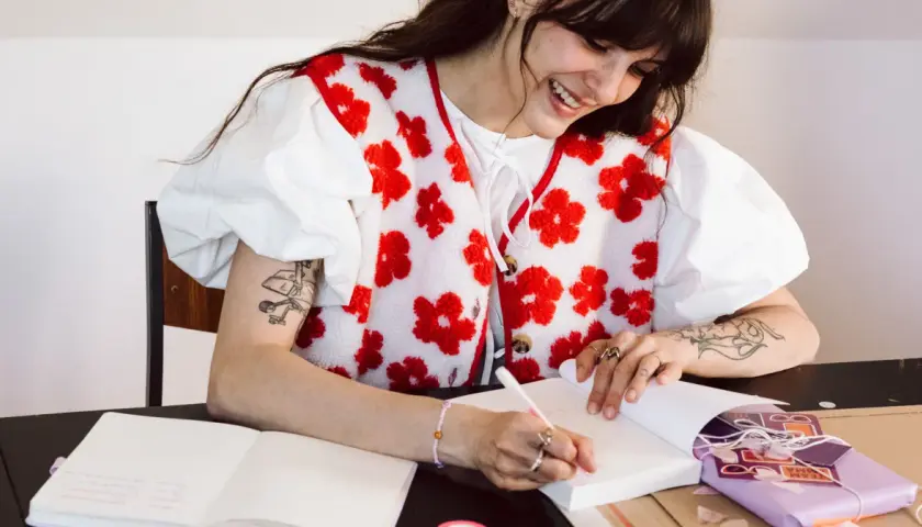 Une jeune femme souriante fait une dédicace livre, stylo à la main, entourée de cadeaux et de carnets.
