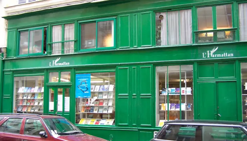 Façade verte d'une librairie L'Harmattan, vitrines remplies de livres. Une voiture rouge et une noire stationnent devant.
