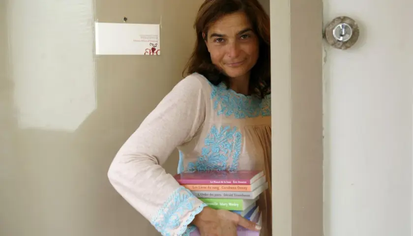 Héloïse d'Ormesson, souriante, tient une pile de livres. Sur le mur, une carte "Éditions Héloïse d'Ormesson".