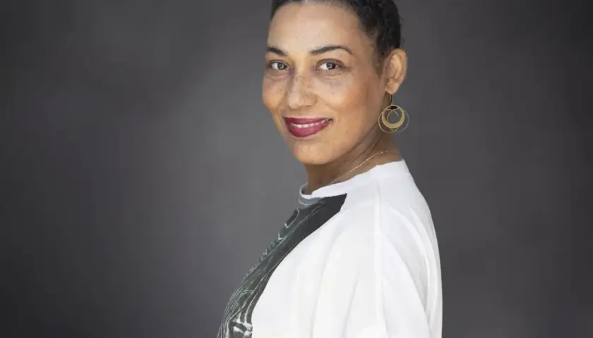 Une autrice au sourire confiant, ses cheveux courts grisonnants encadrant un visage aux taches de rousseur. Elle porte des boucles d'oreilles dorées et un t-shirt blanc.