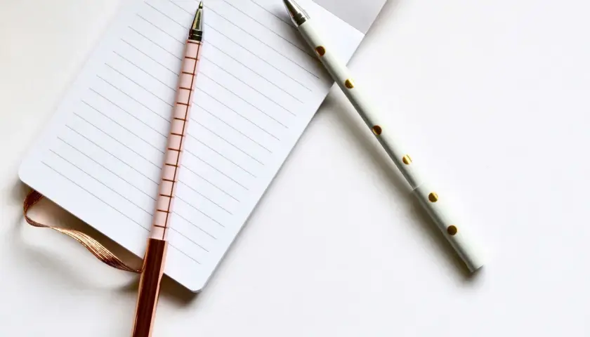 Deux stylos élégants, un rose à carreaux et un blanc à pois dorés, prêts pour l'édition pkj manuscrit sur un carnet ligné.