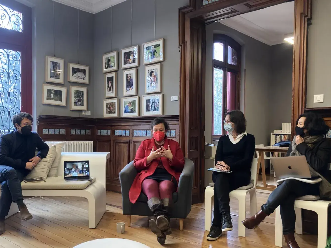 Quatre personnes masquées discutent dans un salon orné de cadres. Une femme en rouge est au centre, semblant raconter une histoire lors d'une résidence d'écriture.