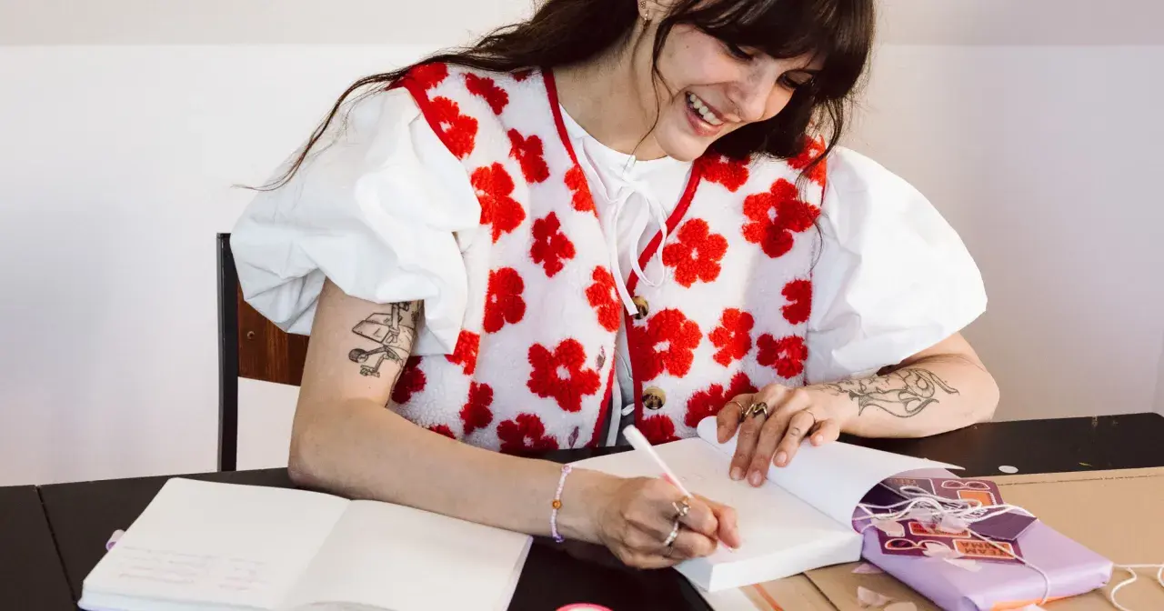 Une jeune femme souriante fait une dédicace livre, stylo à la main, entourée de cadeaux et de carnets.