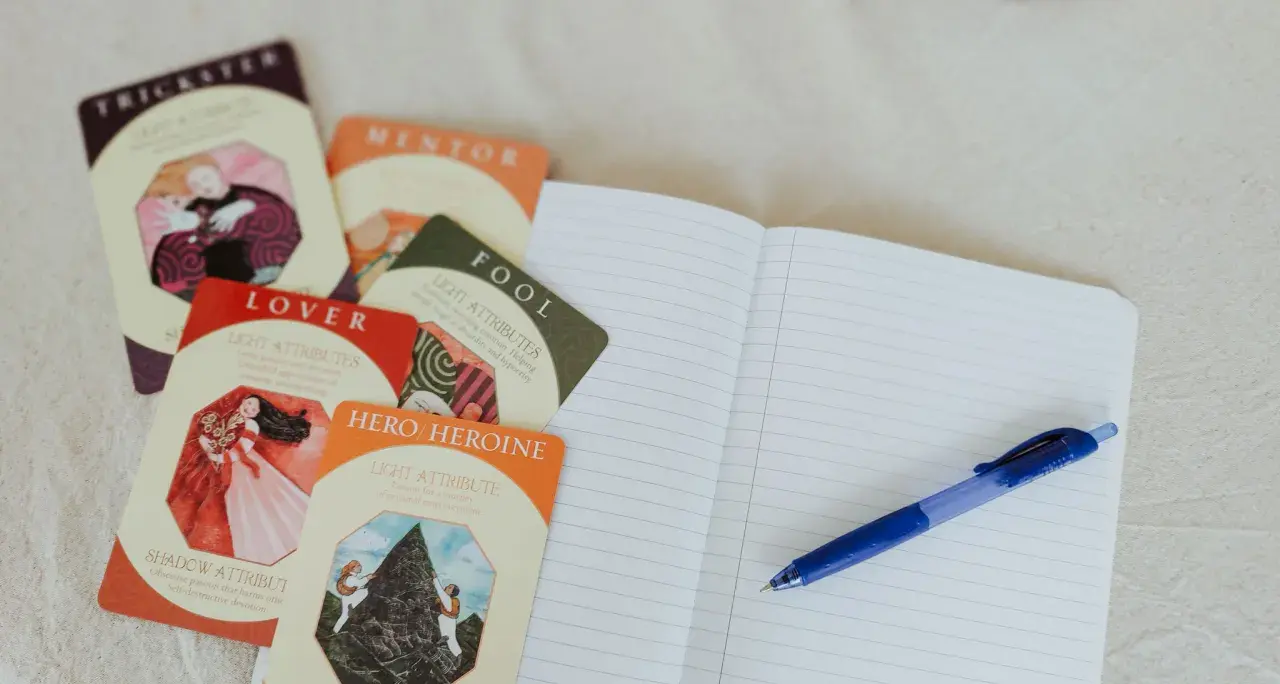 Cartes d'archétypes (Trickster, Lover, Hero/Heroine) et un carnet ouvert avec un stylo, pour une idée de nom pour personnage de roman.