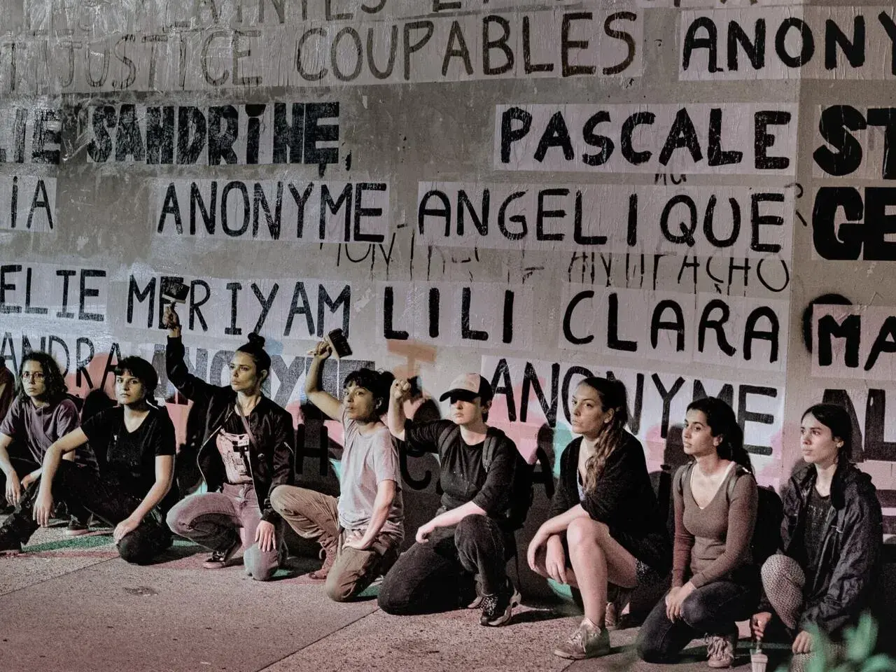 Des femmes s'agenouillent devant un mur couvert de noms, dont "Anonyme", peut-être en soutien à une écrivaine anonyme.