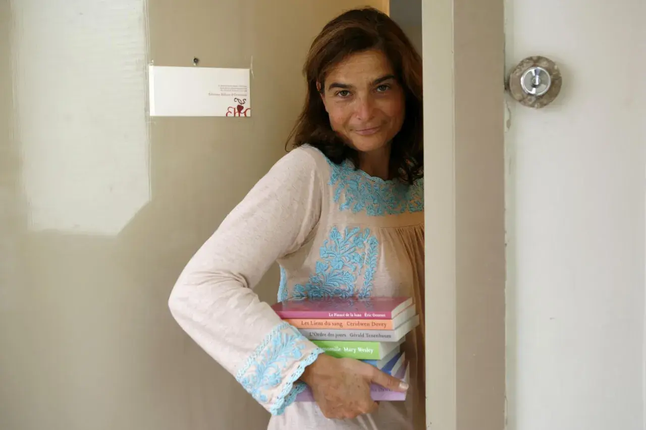 Héloïse d'Ormesson, souriante, tient une pile de livres. Sur le mur, une carte "Éditions Héloïse d'Ormesson".