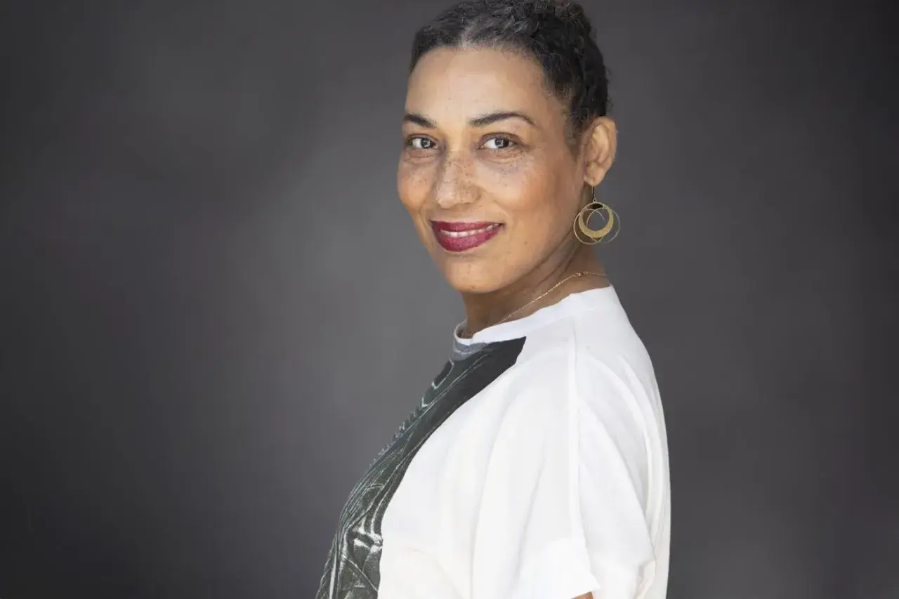 Une autrice au sourire confiant, ses cheveux courts grisonnants encadrant un visage aux taches de rousseur. Elle porte des boucles d'oreilles dorées et un t-shirt blanc.