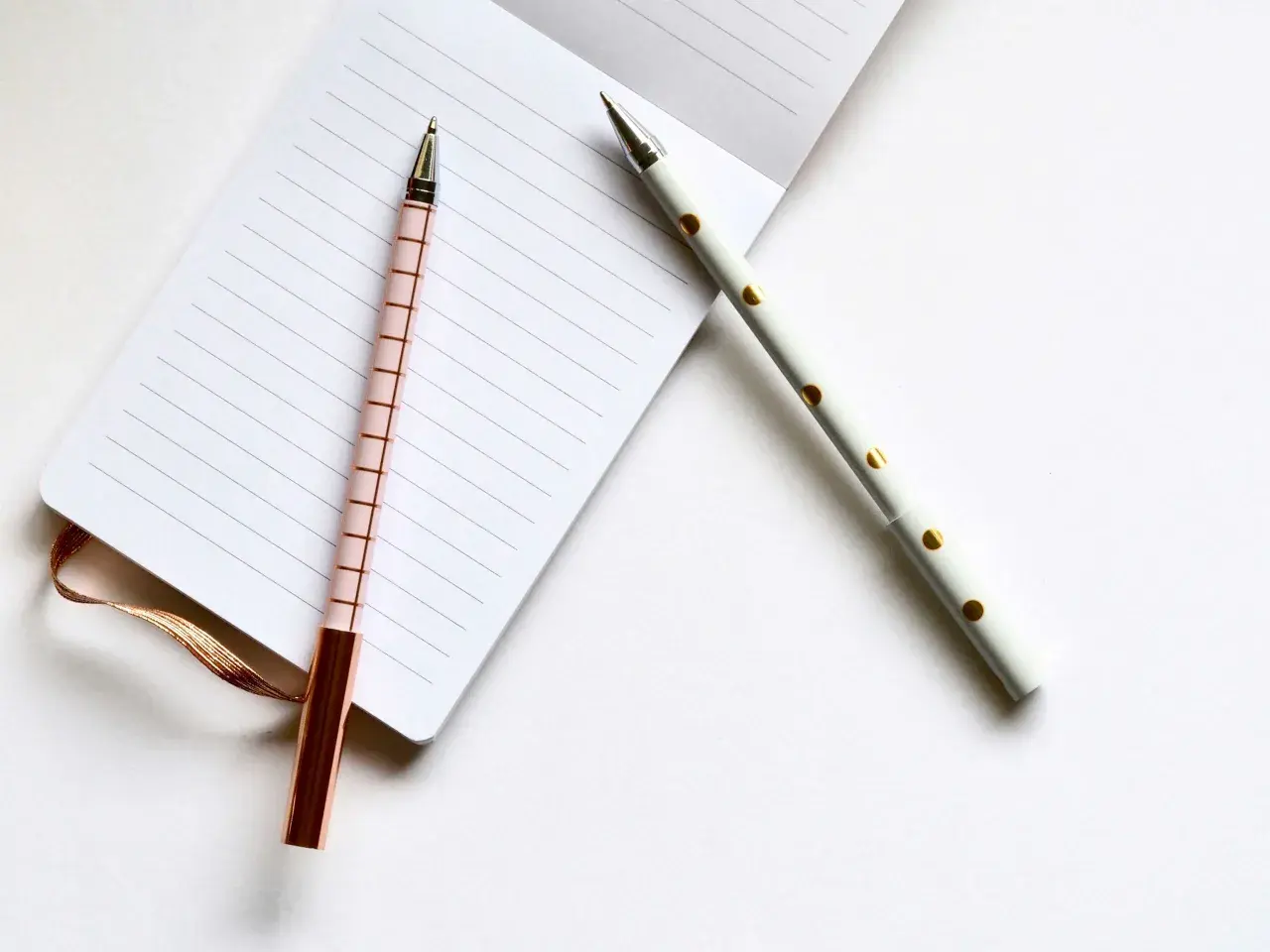 Deux stylos élégants, un rose à carreaux et un blanc à pois dorés, prêts pour l'édition pkj manuscrit sur un carnet ligné.